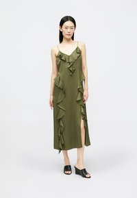 Rochie maxi verde măsliniu cu accente volan în față și fante pe partea laterală. Bretele ajustabile subțiri. Pantofi cu degetul mare negri. Textură netedă.