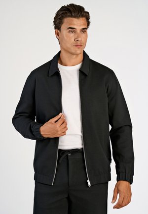 Black zip-up jacket with a collar, made of smooth fabric. Features elastic cuffs and a slim fit. Worn over a white t-shirt.