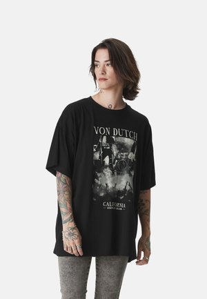 Jeune personne aux cheveux mi-longs et tatouages colorés portant un t-shirt ample noir Von Dutch et un jean gris délavé.
