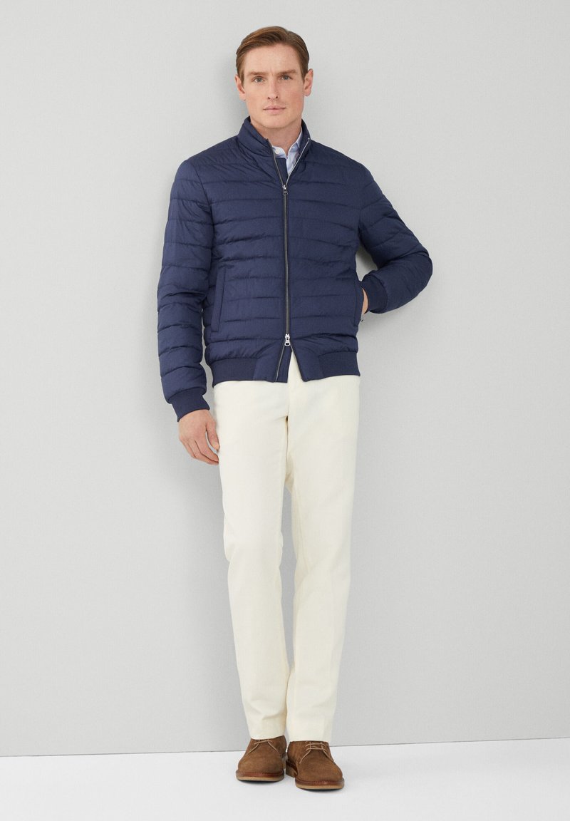 Manteau rembourré matelassé bleu marine avec fermeture éclair et poignets côtelés, associé à un pantalon couleur crème et des chaussures en daim marron.