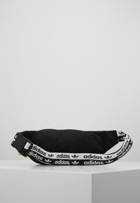 Bolso de cintura negro de Adidas con correa ajustable de marca, que presenta logotipos en blanco y negro y un broche de plástico lateral, exhibido sobre una superficie blanca.