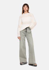 Strickpullover in creme mit offener Maschenstruktur, kombiniert mit hellgrauen, weit geschnittenen Jeans und Slide-Sandalen im Tiermuster.