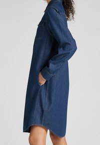 Jeanshemdkleid in Dunkelblau, mit Kragen, langen Ärmeln, seitlichen Taschen und einem geschwungenen Saum. Klassische Nähte sind sichtbar.