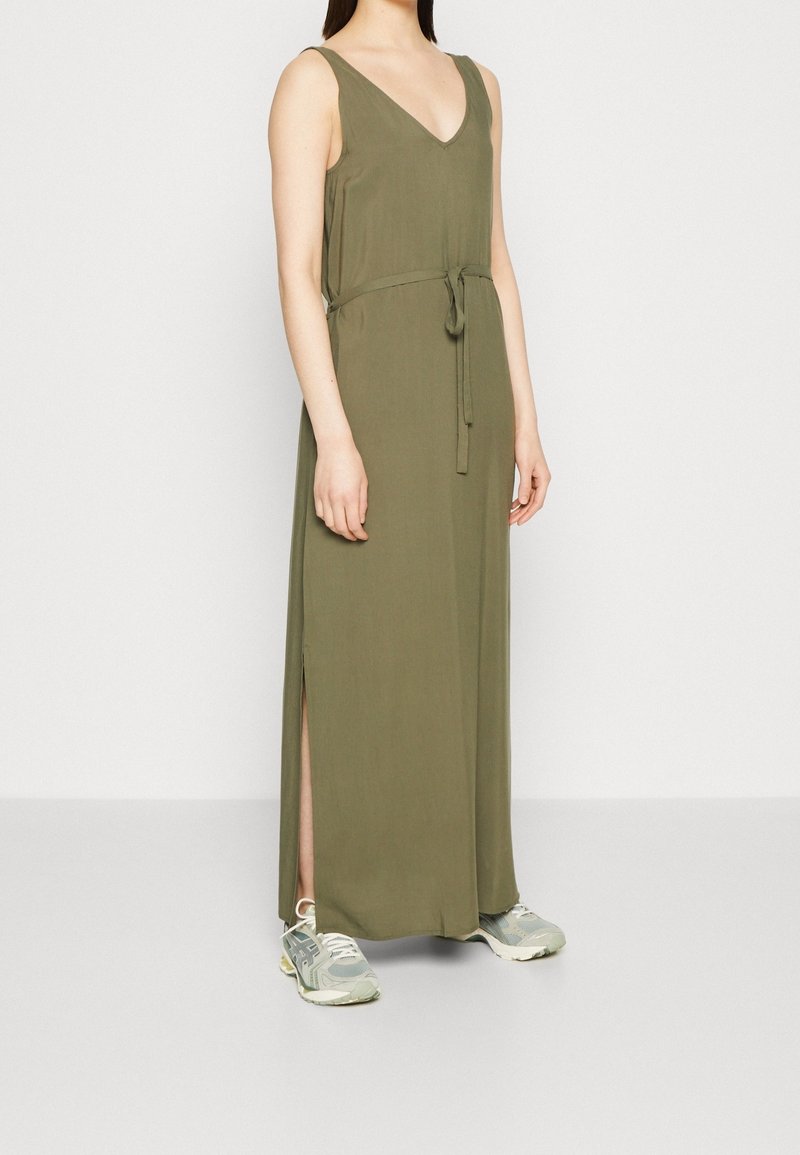 Robe maxi verte olive avec un décolleté en V, sans manches, ceinture ajustable à la taille et fente sur le côté, associée à des baskets grises.