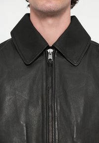 Blouson en cuir noir avec un grand col et une fermeture éclair argentée. La texture est lisse avec un léger éclat, mettant en valeur un savoir-faire de qualité.