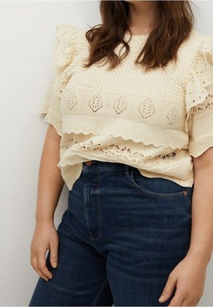 Donna che indossa un maglione di maglia color crema con pizzi e motivi a foglia, abbinato a jeans a vita alta blu scuro, in piedi davanti a uno sfondo neutro.