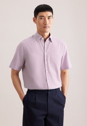 Uomo che indossa una camicia bianca a maniche corte con motivo a quadri rossi e pantaloni blu navy scuro, in piedi con le mani nelle tasche davanti a uno sfondo semplice.