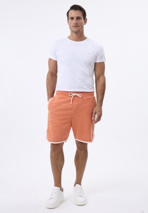 Shorts deportivos naranjas con cordón blanco y rayas laterales, combinados con una camiseta blanca sencilla y zapatillas blancas. Diseño simple y atlético.