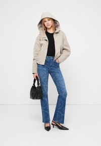 Μπεζ ζακέτα με κουκούλα, μαύρο τοπ, μπλε καμπάνα τζιν με σχέδια, μαύρη τσάντα και μαύρα μυτερά παπούτσια. Απλός σχεδιασμός με casual εμφάνιση.