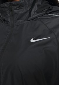Czarna kurtka Nike z zapięciem na zamek, wysokim kołnierzem i eleganckim designem. Posiada wyraźne białe logo Nike na lewej piersi.