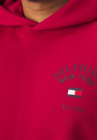 Červená mikina s kapucí vyrobená z bavlněné směsi. V přední části je grafický text "HILFIGER NEW YORK" a design vlajky v bílé, modré a červené barvě.
