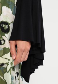 Handen vilar vid grön blommig tyg och lager av svart ärm med volanger, mot en enkel ljus bakgrund.