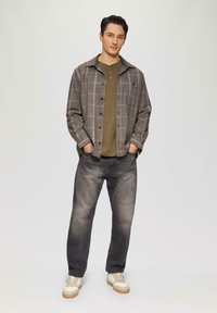 Camicia a quadri grigia con bottoni sopra una maglietta henley verde oliva, abbinata a jeans scuri e sneakers beige. Texture morbide, design casual.
