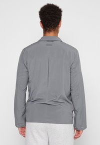 Leichte graue Jacke mit Rückenbelüftung und einfachem Kragen. Hergestellt aus glattem Stoff mit Druckknöpfen an den Bündchen.