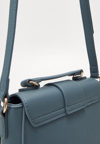 Blå läderhandväska med en strukturerad design, som har ett handtag upptill och en justerbar rem, accentuerad med guldf ärgade detaljer och sömmar.