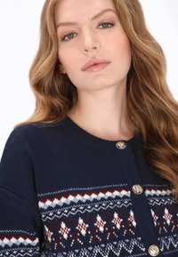 Cardigan bleumarin cu un model geometric în alb, roșu și albastru, cu nasturi aurii și guler rotund. Țesătură moale și texturată din tricot.