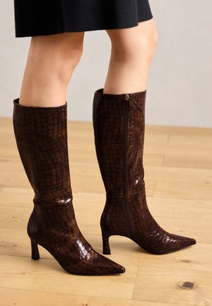 Bruine laarzen tot de knie met een krokodillenstructuur, puntige neus en een lage hak. Voorzien van een zijrits voor sluiting en een glanzende afwerking.