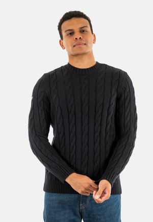 Man in een zwart kabelgebreid sweater en blauwe jeans, die de mouw aanpast, staande tegen een effen lichte achtergrond.