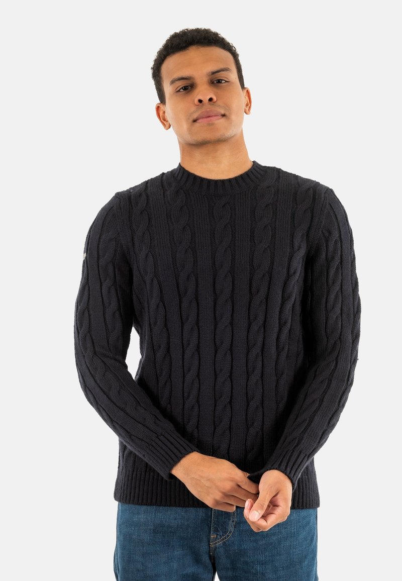 Man in een zwart kabelgebreid sweater en blauwe jeans, die de mouw aanpast, staande tegen een effen lichte achtergrond.