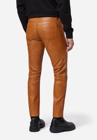 Bruine leren slim-fit broek met een gladde textuur, met achterzakken, subtiele stikseldetails en gedragen met zwarte enkellaarzen.