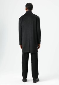 Zwart lange jas met een gladde textuur, enkele inkeping aan de achterkant, gecombineerd met zwarte broek met wijde pijpen en zwarte schoenen.