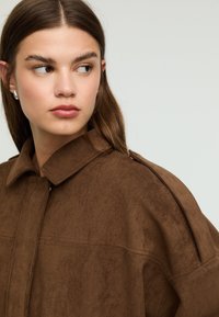 Giacca in suede marrone con scollo a collo, caratterizzata da una texture liscia e un dettaglio di bottoni laterali, mostrata su uno sfondo neutro.