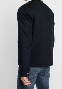 Svart bomulljacka med ett fårskinnskrage, med en åtsittande design, knappdetaljer och en slät textur. Synlig sömdetalj.