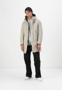 Beige polstret frakk med lynlås og knaplukning, har en hætte, sidelommer og et logo-patch på skulderen. Båret over en lys skjorte.