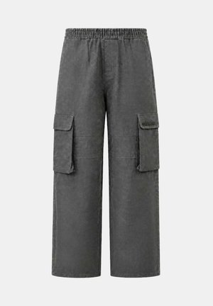 Un hombre con gafas de sol, una sudadera de cuadros coloridos y pantalones cargo grises sostiene una mochila gris y negra en un estudio.
