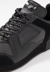 Baskets noires en cuir texturé et panneaux en mesh, lacets noirs et blancs à motifs, et languette marquée avec œillets métalliques.
