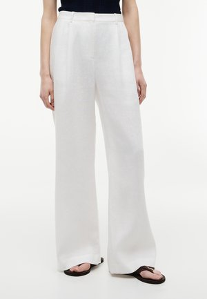 Mujer vistiendo pantalones blancos de tiro alto y pierna ancha con presillas para cinturón, combinados con top negro y sandalias negras.