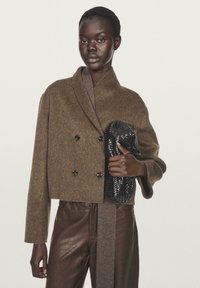 Donna di pelle scura che indossa un giubbotto doppiopetto marrone in lana e pantaloni di pelle, mentre tiene una clutch intrecciata nera su uno sfondo semplice.