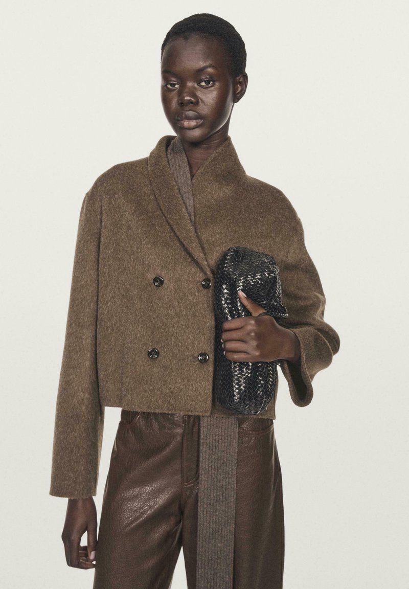 Donna di pelle scura che indossa un giubbotto doppiopetto marrone in lana e pantaloni di pelle, mentre tiene una clutch intrecciata nera su uno sfondo semplice.