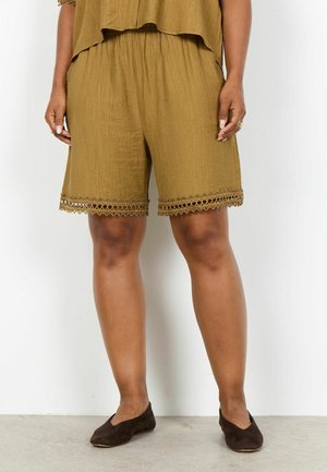 Brune teksturerede shorts med blonderkant, båret af en person, der står, parret med mørkebrune slip-on sko på et lyst gulv.