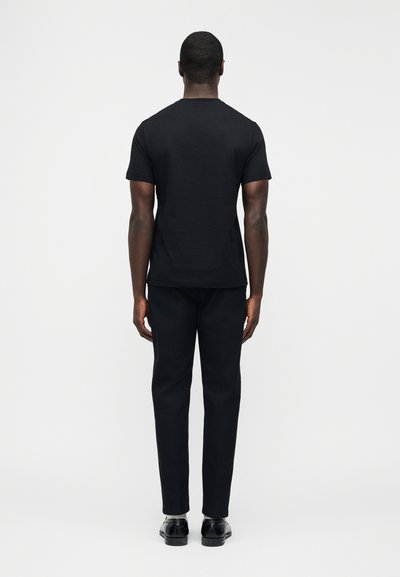 Ενήλικας που στέκεται με την πλάτη, φοράει απλό μαύρο κοντομάνικο μπλουζάκι, μαύρο παντελόνι, λευκές κάλτσες και μαύρα επίσημα παπούτσια, με λευκό φόντο.