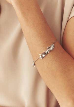 Bras portant un délicat bracelet en argent avec plusieurs petits charms en forme de feuille sur un fond de tissu beige.