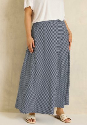 Frau trägt ein weißes Top, einen marineblau-weiß gestreiften langen Rock und beige flache Sandalen, steht vor einem schlichten beigen Hintergrund.