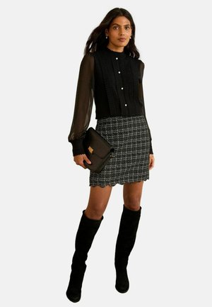 Czarna bluzka z przezroczystymi rękawami i zapięciem na guziki, w połączeniu z teksturowaną spódnicą w czarno-białą kratę. Modelka trzyma czarną torebkę.