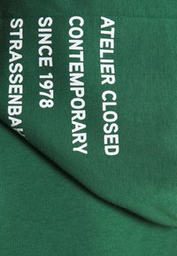 Tessuto verde con testo stampato in bianco recante le parole "ATELIER CHIUSO," "CONTEMPORANEO," "DAL 1978," e "STRASSENBAHN." Texture liscia.