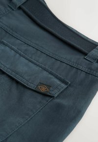 Tissu bleu foncé avec un rabat de poche cousu, comportant une petite étiquette noire avec un logo trident orange, reposant sur une surface blanche.