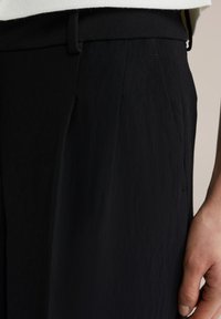 Pantalonii negri, texturizați, au un design pliat și un talie netedă. Prim-planul arată detalii ale țesăturii și o mână odihnindu-se pe lateral.