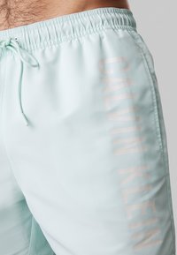 Pantaloni da bagno azzurri chiari con finitura texturizzata, dotati di vita con cordoncino e logo "CALVIN KLEIN" in un colore chiaro lungo il lato.