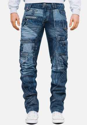 Hombre con vaqueros patchwork azules con varios bolsillos, detalles de costuras y zapatillas blancas, de pie con los brazos relajados a los lados.