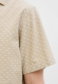 Camicia a maniche corte in tessuto beige chiaro con un motivo testurizzato di piccoli simboli bianchi. Presenta un colletto classico e una vestibilità rilassata.