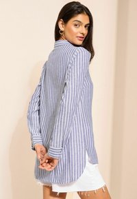 Kék-fehér csíkos, hosszú ujjú ing magas-alacsony szegéllyel és gombos mandzsettával, könnyű anyagból készült. Fehér, rojtos szoknyával viselve.