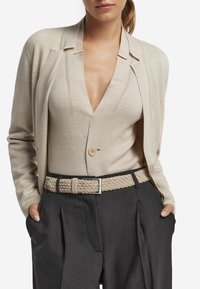 Beige stickad cardigan med djup V-ringning, som matchas med en beige knäppt topp och grå högmidjade byxor, prydda med ett flätat beige bälte.
