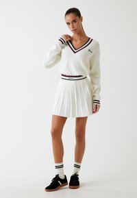 Suéter blanco de punto con escote en V y rayas en burdeos y azul marino. Falda blanca plisada con una cinturilla de marca. Zapatillas negras con detalles en blanco.