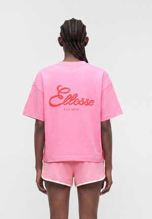 Henkilö, jolla on punotut hiukset, pukeutuneena vaaleanpunaiseen lyhythihaiseen paitaan ja yhteensopiviin shortseihin, paidan takana näkyy punainen "Ellesse EST 1959" -logo.