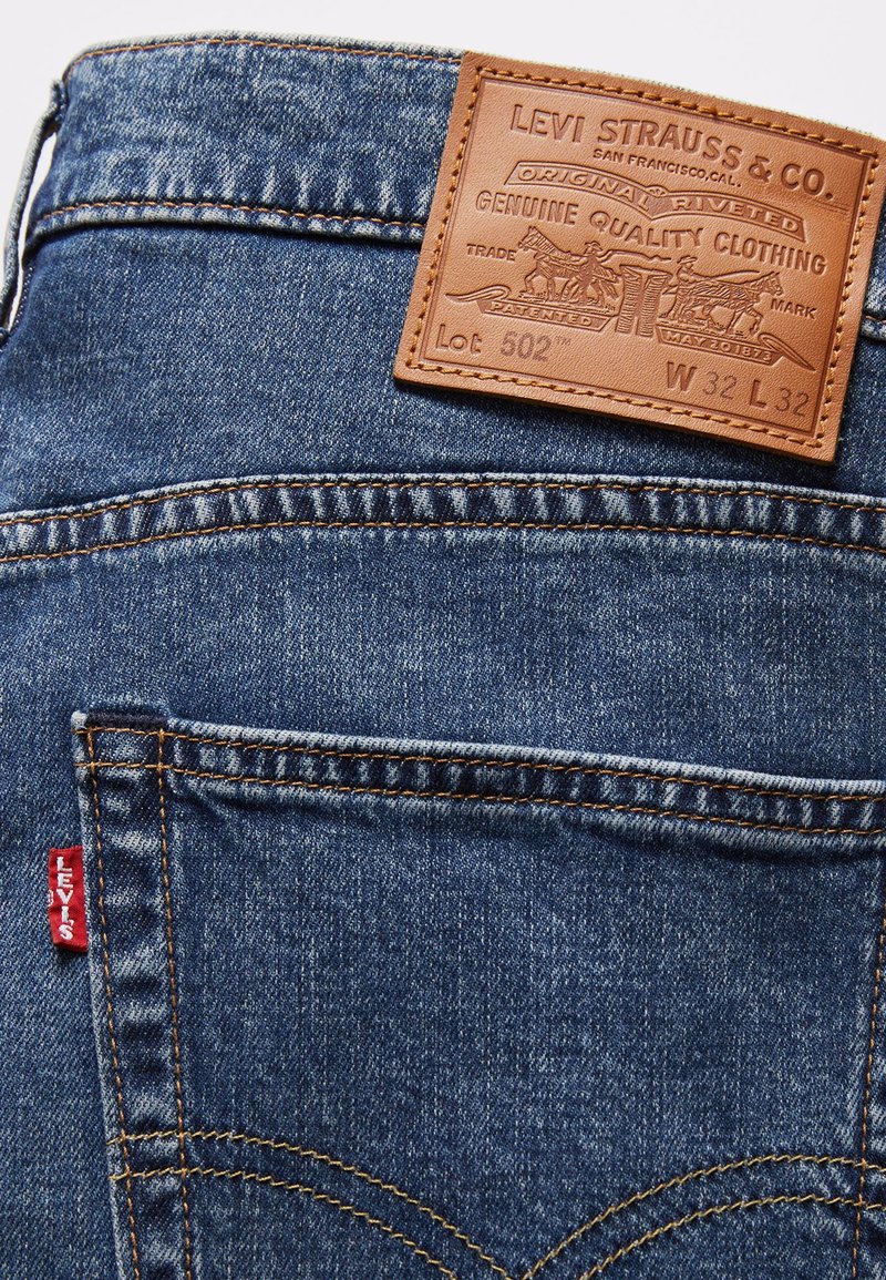 Die Levi's 502 Jeans aus dunkelblauem Denim verfügen über ein Lederpatch mit eingeprägtem Logo und Stickdetails, einschließlich eines roten Tabs an der Seite.