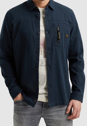 Homme portant une chemise bleu foncé déboutonnée sur un t-shirt blanc graphique et un jean gris, les mains dans les poches.